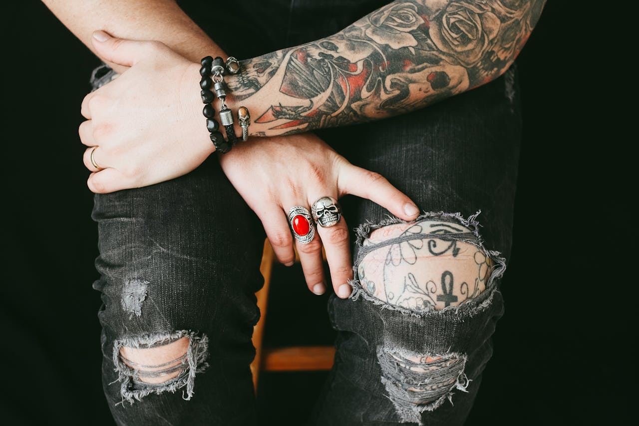 about-02 Close-up of tattooed hands with rings and bracelets over ripped jeans, showcasing fashion and style.