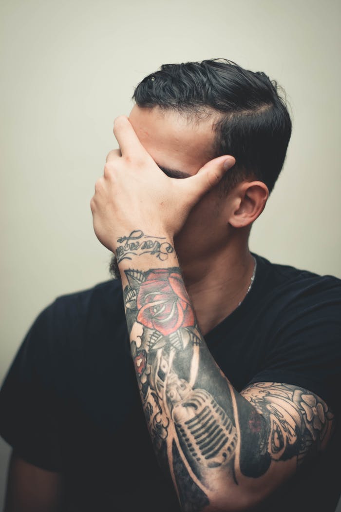 menu-01 Stylish man with detailed arm tattoos covers face with hand, showcasing intricate body art.