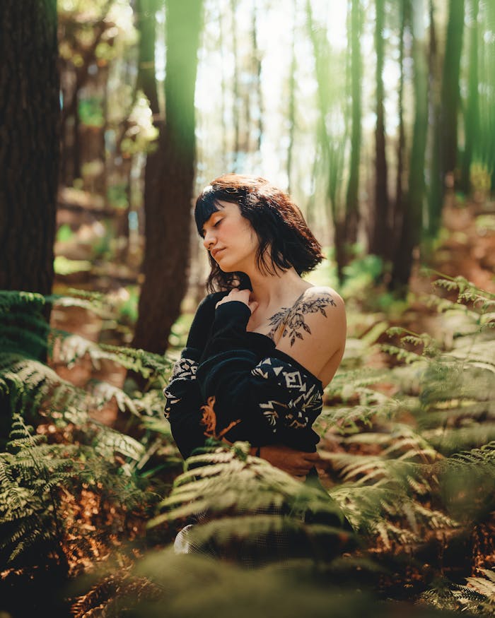 menu-11 A serene woman with a tattoo enjoys nature in a sunlit, fern-lined forest.