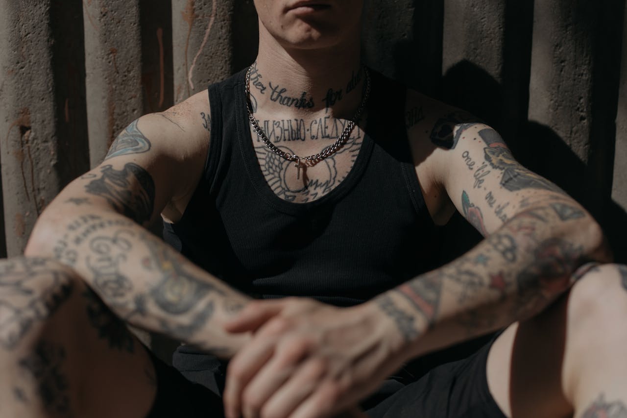 about-02 Close-up of a tattooed person in a black tank top sitting against a corrugated wall in moody lighting.
