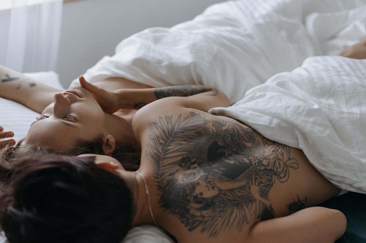 about-04 An intimate moment of a tattooed couple lying together on a bed with white linens.