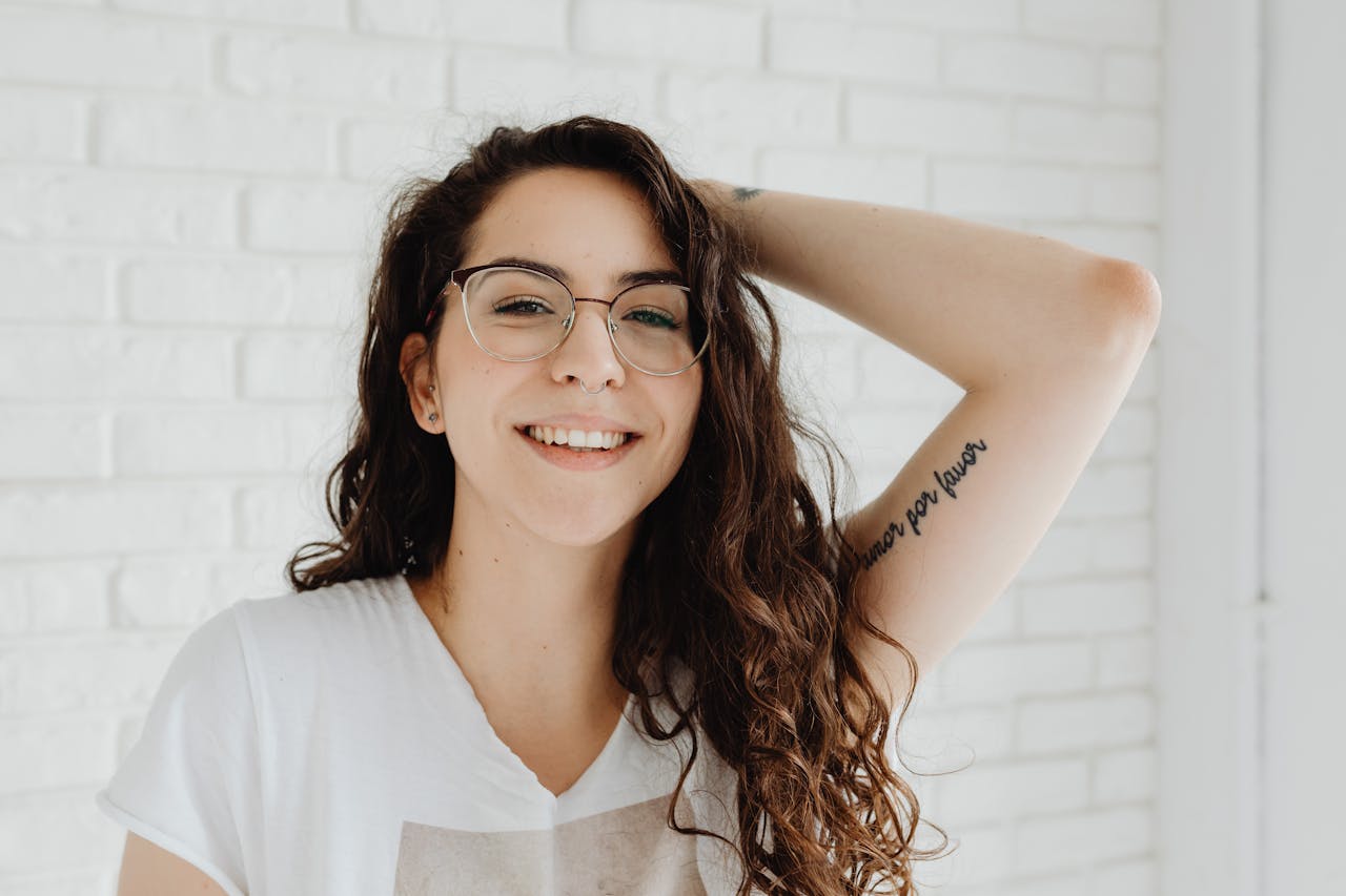 menu-02 Portrait of a young woman smiling with eyeglasses and a tattoo, wearing a white shirt.